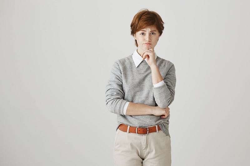 A young woman with short red hair stands with her arms crossed, with a concerned expression, resting her hand on her chin.