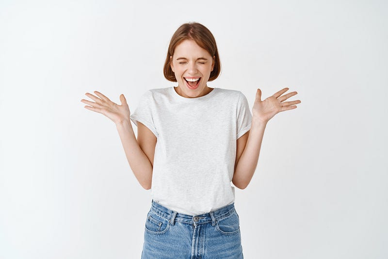 A young woman with strawberry blond hair hold her hands up in excitement with her eyes closed tightly, her mouth open and smiling.