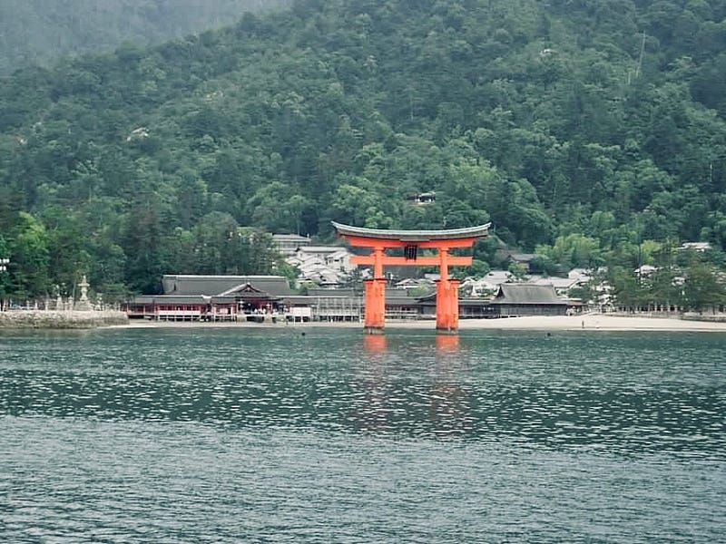 Orange temple gates rising out of water in front of a Shinto temple and forested mountain.