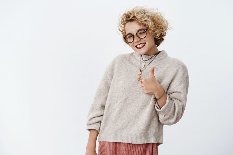 A young woman with short curly blonde hair and glasses gives an enthusiastic thumbs up with a wink.