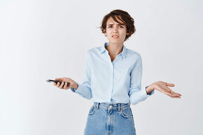 A young woman with short dark brown hair shrugs her shoulders and looks both confused and disgusted while holding a mobile ph