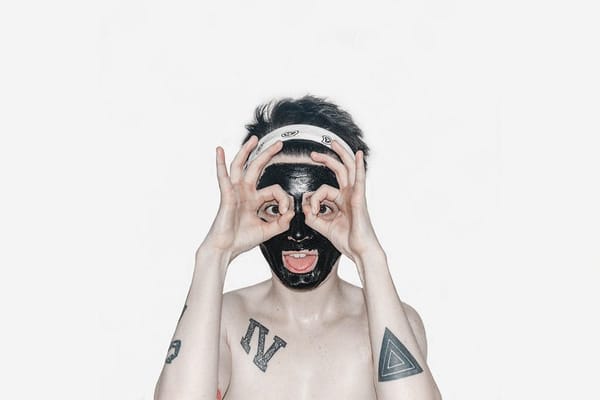 Photo of a light-skinned person with short hair and several tattoos wearing a mud facial mask and making a silly face.